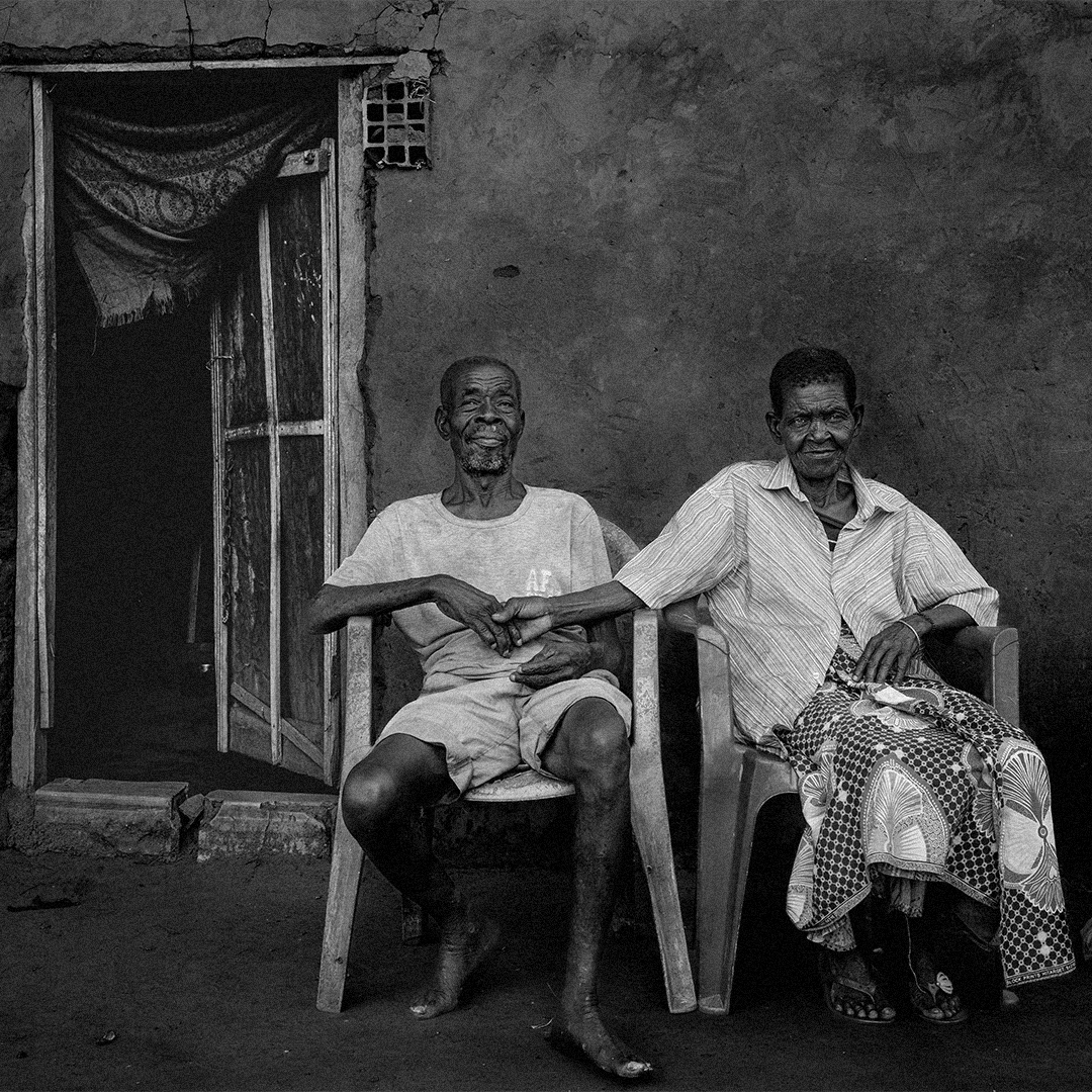 Casal idoso sentado à porta de casa, segurando-se pela mão com expressão de cumplicidade.