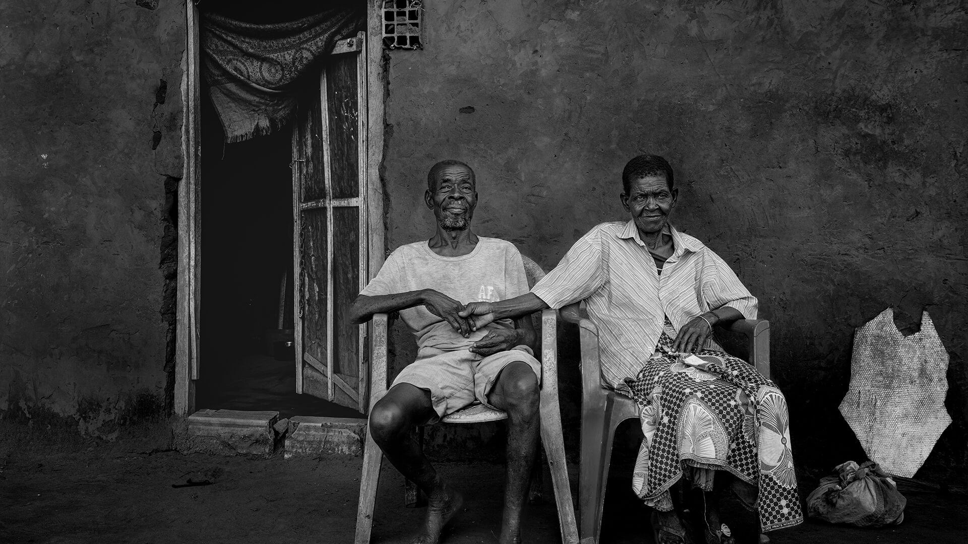 Casal idoso sentado à porta de casa, segurando-se pela mão com expressão de cumplicidade.