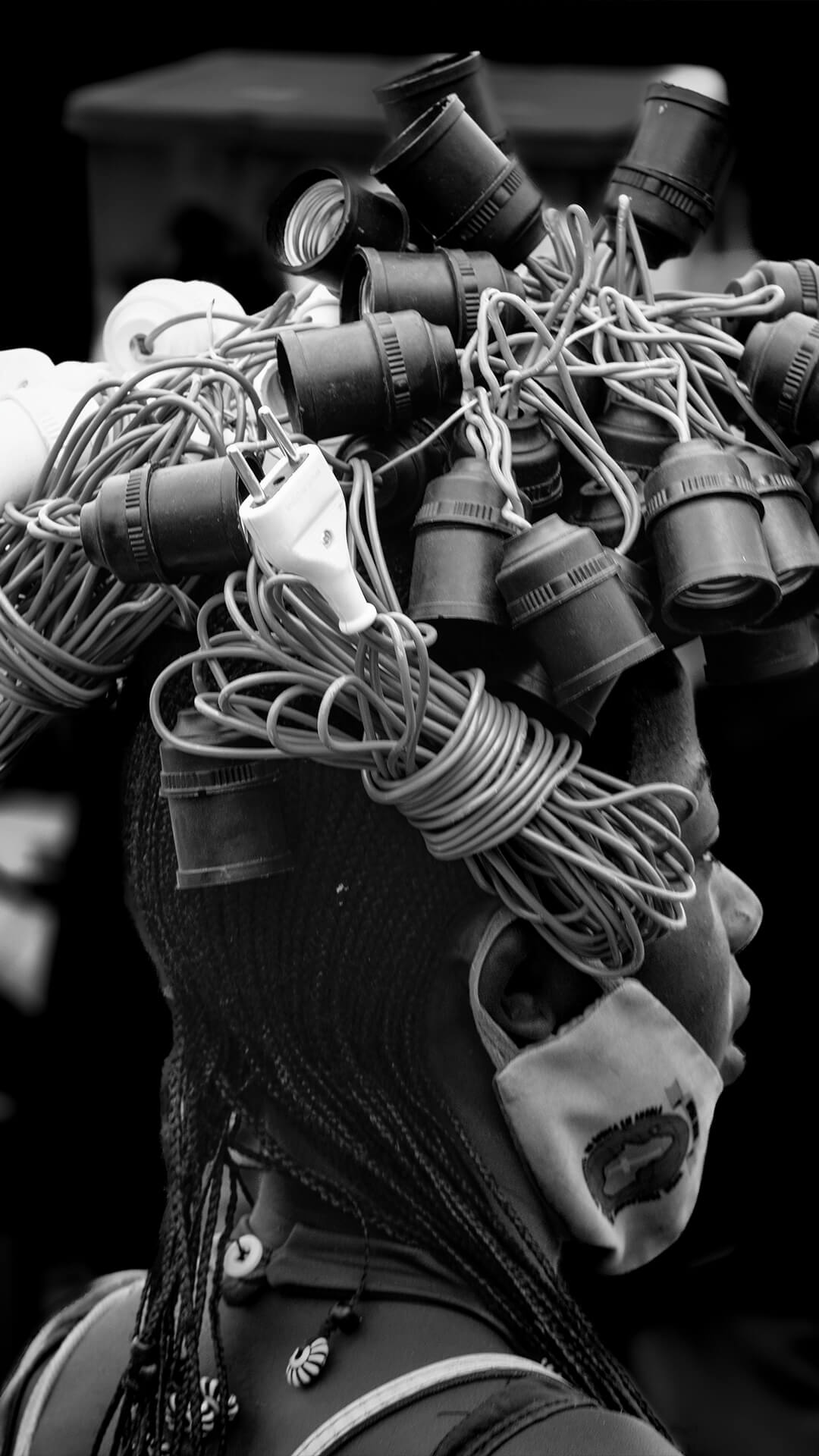 Fotografia artística em preto e branco de uma mulher Himba com adereços tradicionais em perfil, destacando o tema 'Traços de [es]culturas'.